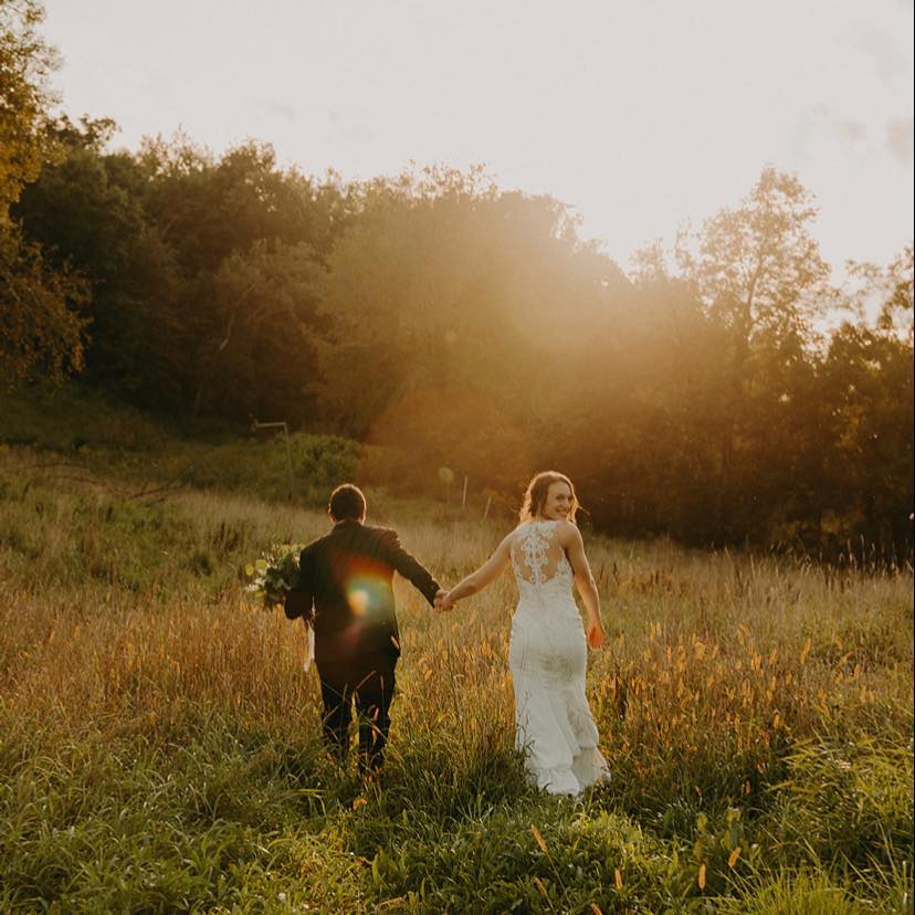 a bride and groom walk hand in hand in a field at sunset.