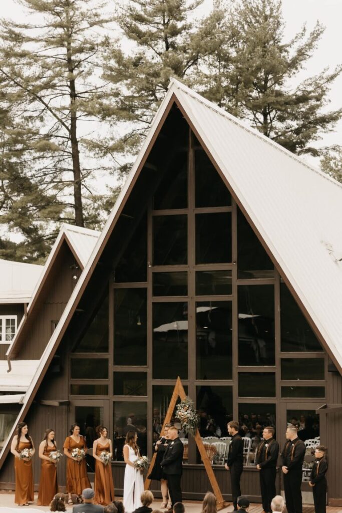 A wedding party with orange dresses and dark suits stands up during the ceremony in front of the lodge at Mt. La Crosse.