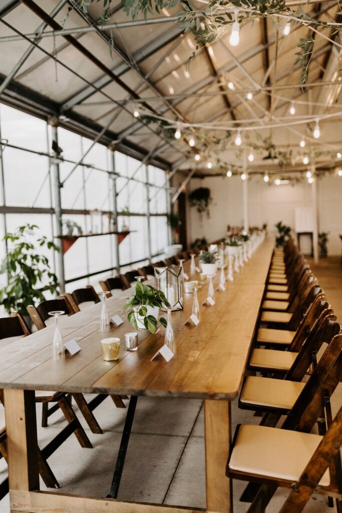 A long wooden table and chairs set with champagne glasses, place cards, and plants for a wedding reception at The Greenhouse At Bittersweet.